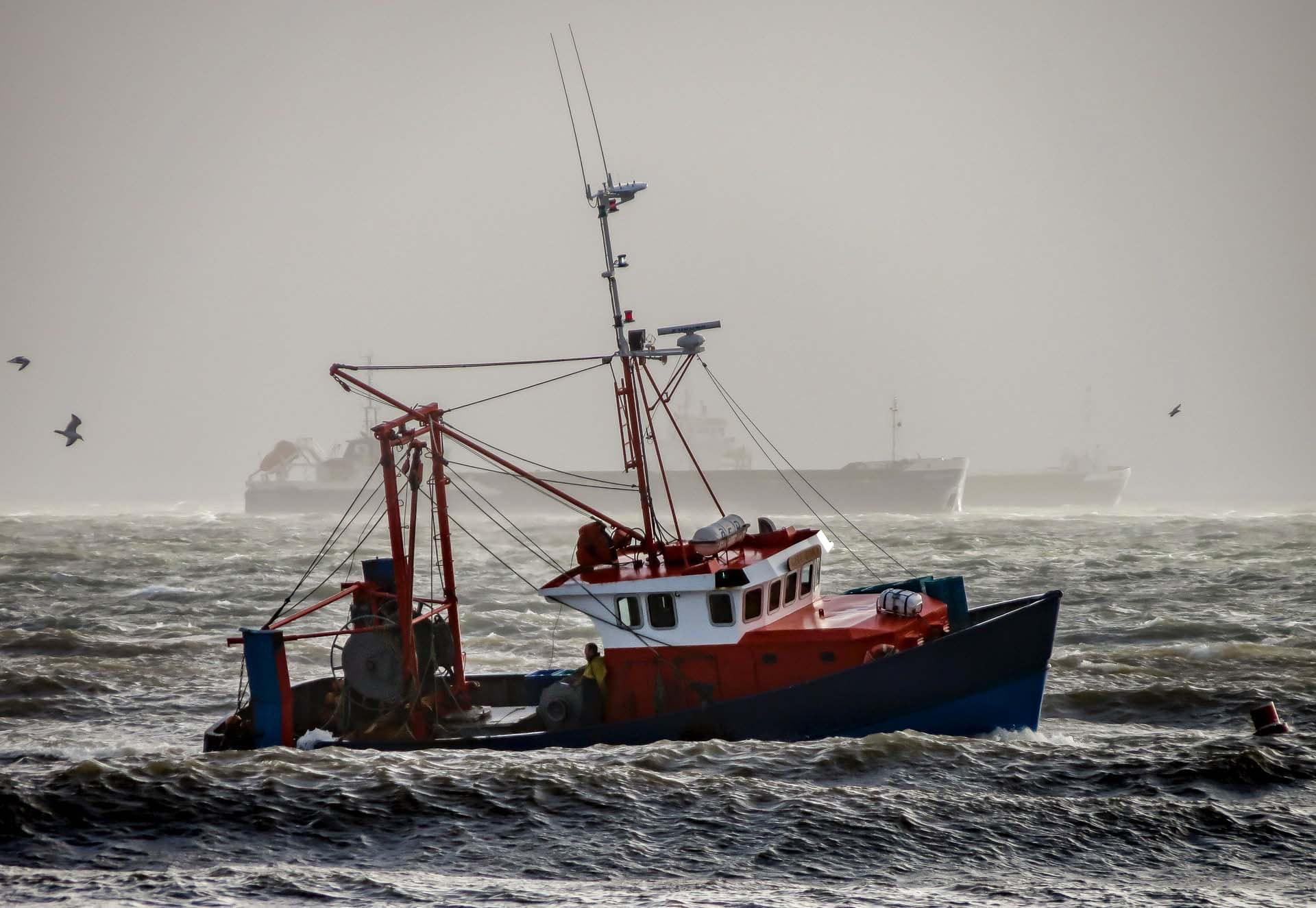 Commercial fishing trawler operating in rough coastal seas with other vessels visible in misty background.