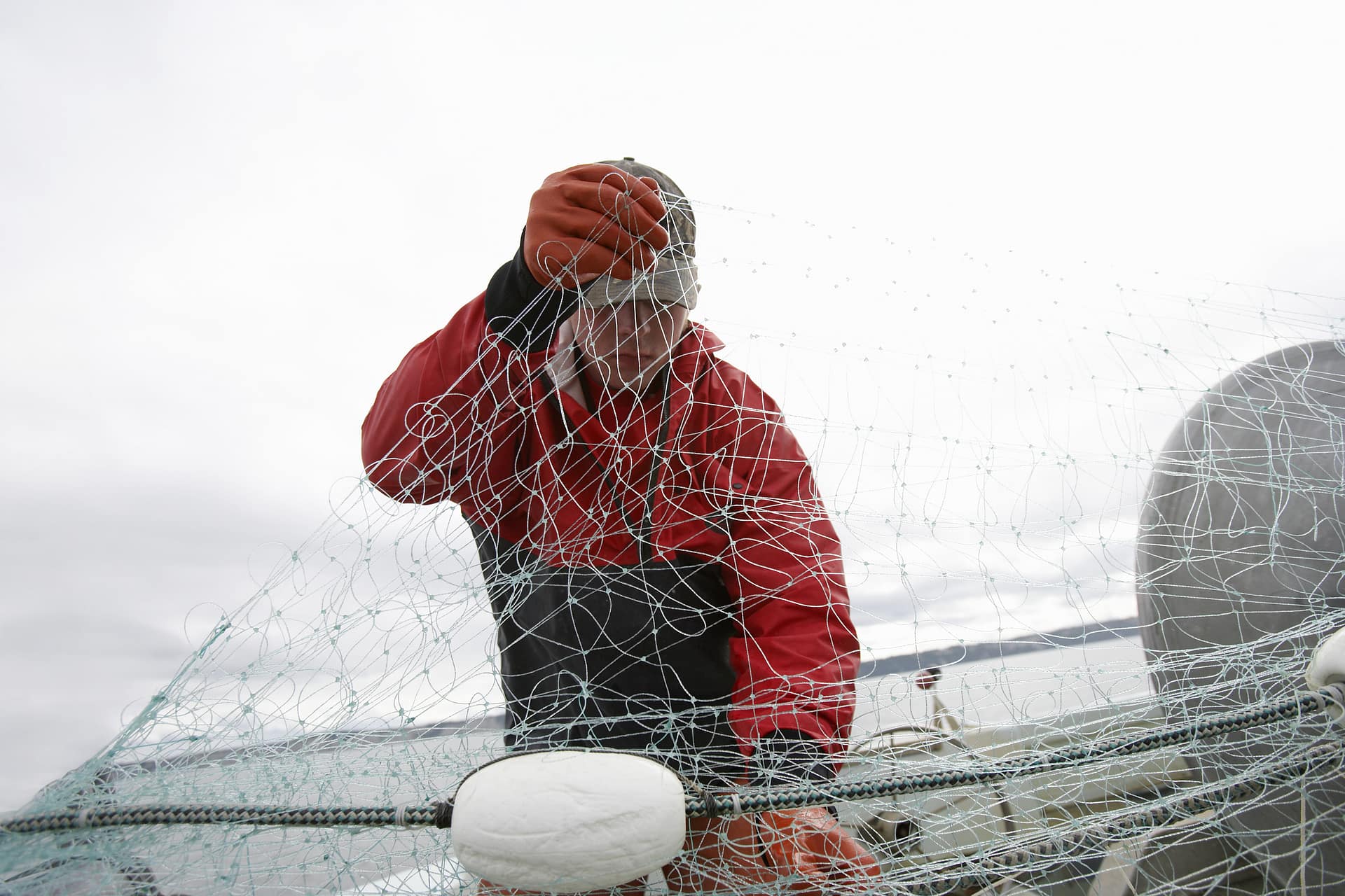 A deckhand in a red jacket and gloves inspecting a tangle of fishing net on the deck of a commercial vessel under an overcast sky.