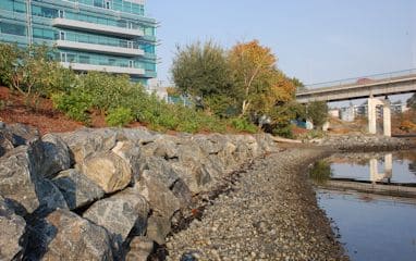 Rock armored shoreline and narrow gravel beach in front of a modern glass building and bridge over calm still water.
